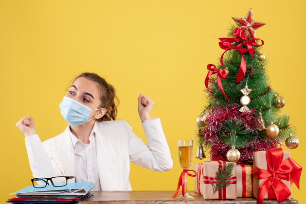 工作正面图女医生戴着无菌口罩坐在黄色背景上 带着圣诞树和礼品盒成人实验室背景