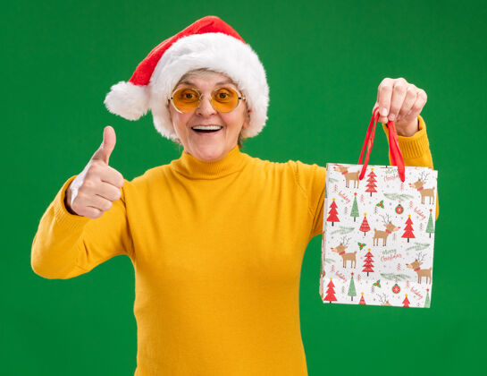 背景一位戴着太阳眼镜 戴着圣诞帽的老妇人手持纸质礼品袋 竖起大拇指 在绿色的背景上 留有复印空间 给人留下深刻印象印象深刻快乐圣诞快乐