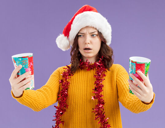 快乐困惑的年轻斯拉夫女孩戴着圣诞帽 脖子上戴着花环 手里拿着纸杯 看着紫色背景上的纸杯 还有复印空间看困惑围着