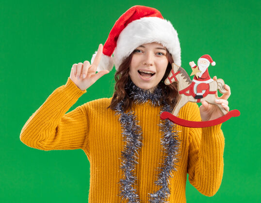 花环快乐的年轻斯拉夫女孩 戴着圣诞帽 脖子上戴着花环 把圣诞老人抱在摇马装饰上 在绿色背景上用复制空间向上指脖子摇摆帽子