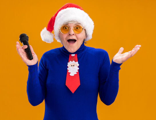 眼镜戴着太阳眼镜 戴着圣诞帽 系着圣诞领带 拿着麦克风的惊讶的老妇人被隔离在橙色的背景上 还有复制空间圣诞快乐年麦克风