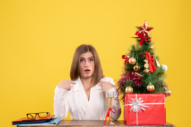 前面前视图女医生坐在桌子前 带着圣诞礼物和黄色背景的树人生活女医生