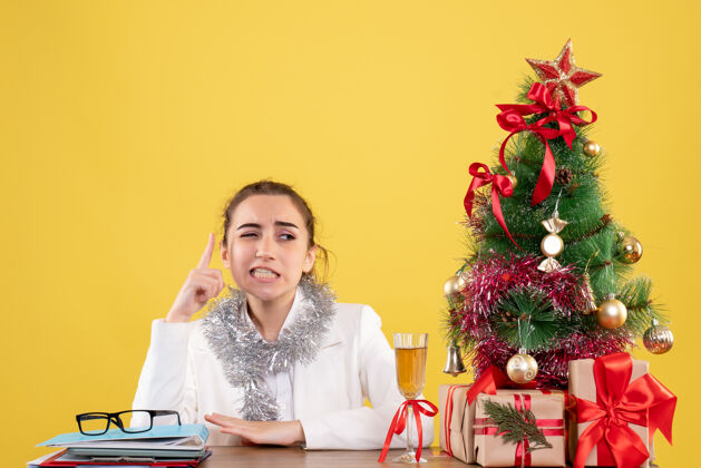 桌子前视图：女医生坐在桌子后面 拿着圣诞礼物和黄色背景上的树女性前面圣诞树