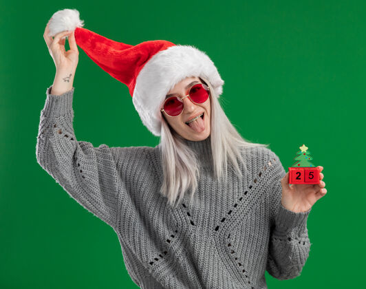 日期年轻的金发女郎穿着冬季毛衣 戴着圣诞老人帽 戴着红眼镜 手里拿着玩具方块 上面写着圣诞节的日期 站在绿色的背景上开心地伸出舌头坚持眼镜毛衣