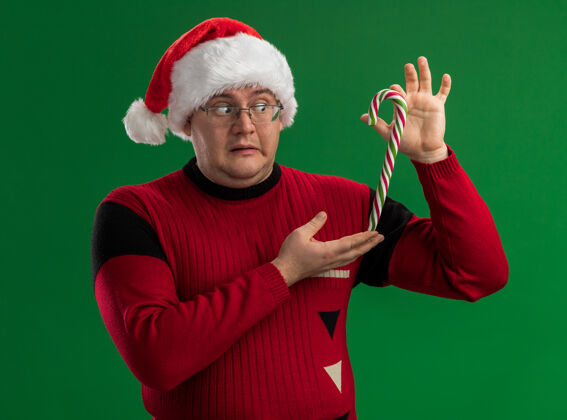 糖果令人印象深刻的成年男子戴着眼镜和圣诞帽手持圣诞甜手杖垂直看着它孤立的绿色背景圣诞老人垂直成人