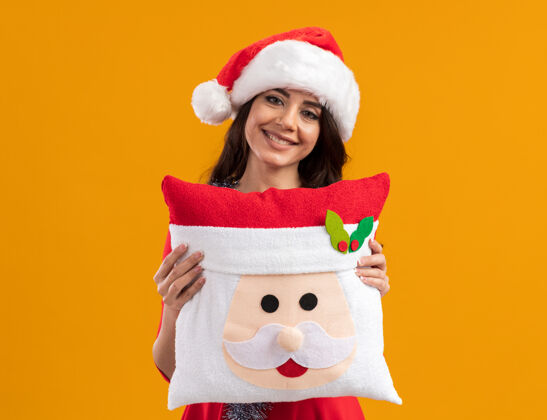 枕头微笑的年轻漂亮女孩戴着圣诞帽 脖子上戴着金箔花环 抱着圣诞老人的枕头看橙色帽子微笑