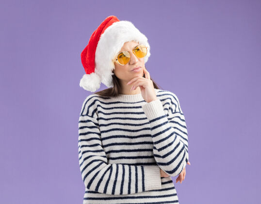 帽子体贴的年轻白种女孩戴着太阳眼镜 戴着圣诞帽 把手放在下巴上 看着侧面圣诞快乐紫色下巴