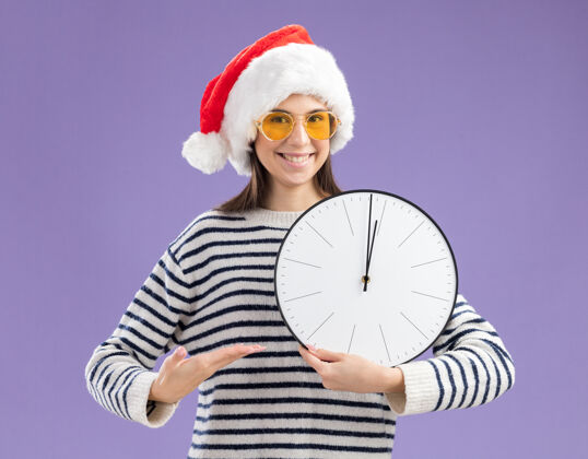 新戴着太阳眼镜 戴着圣诞帽 用手指着时钟的年轻高加索女孩年轻指向圣诞快乐