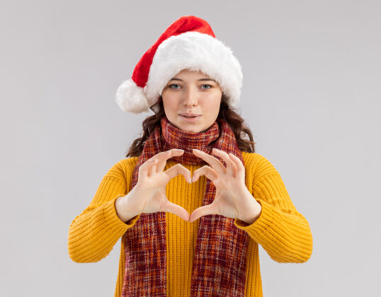 新快乐的年轻斯拉夫女孩戴着圣诞帽 脖子上围着围巾 打着心形手势圣诞圣诞快乐圣诞老人