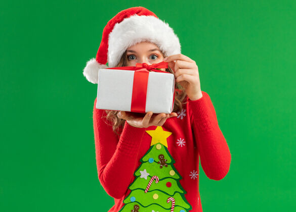拿着身穿红色圣诞毛衣 戴圣诞帽的年轻女子拿着礼物准备打开礼物 站在绿色的背景下 她很感兴趣好奇礼物年轻