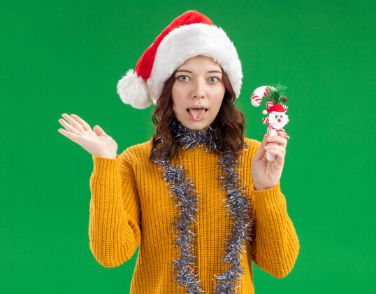 圣诞老人兴奋的年轻斯拉夫女孩 戴着圣诞帽 脖子上戴着花环 手里拿着糖果手杖 手一直张开复制脖子开放