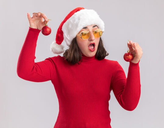 穿着穿着红色毛衣 戴着圣诞帽 戴着眼镜 拿着圣诞球的年轻女孩站在白色的墙上 困惑地看着他们 试图做出选择毛衣制造圣诞节