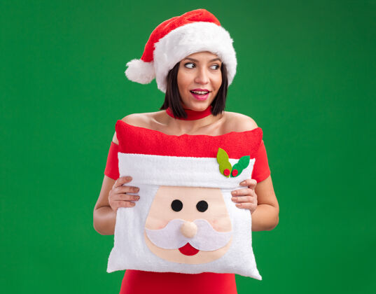 圣诞老人快乐的年轻女孩戴着圣诞帽 抱着圣诞老人的枕头 看着隔离在绿墙上的一面戴侧快乐