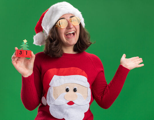 立方体穿着圣诞毛衣的小女孩戴着圣诞帽戴着眼镜拿着玩具方块和新年约会快乐快乐地举起手臂站在绿色的墙上穿着站着毛衣