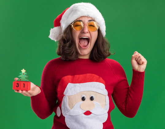 日期穿着圣诞毛衣的小女孩戴着圣诞帽戴着眼镜拿着玩具立方体和新年约会疯狂的快乐和兴奋的握紧拳头站在绿色的墙上拳头站立圣诞节