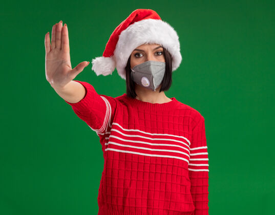 年轻严肃的年轻女孩戴着圣诞帽和防护面具在绿色的墙上做停止手势停止穿手势