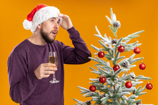 圣诞节一个穿着紫色毛衣 戴着圣诞帽的年轻人 手里拿着一杯香槟 站在圣诞树旁 隔着橘色的墙疑惑地抬头望着玻璃杯男人帽子