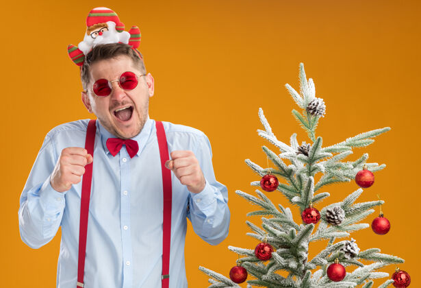 蝴蝶结戴着吊带领结 戴着圣诞老人和红眼镜的年轻人站在圣诞树旁 在橘色的墙上快乐而兴奋穿着圣诞老人轮辋
