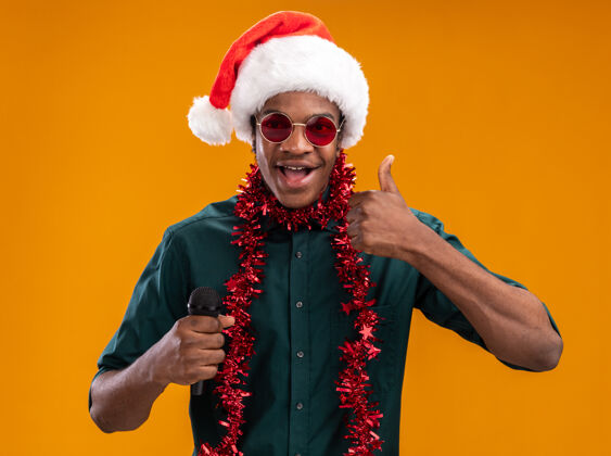 展示戴着圣诞帽戴着花环戴着眼镜拿着麦克风微笑着竖起大拇指站在橙色的墙上的非裔美国人站立男人拇指