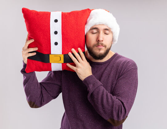 男人一个穿着紫色毛衣 戴着圣诞帽的年轻人抱着一个有趣的枕头 闭着眼睛站在白色的墙上毛衣关闭站着