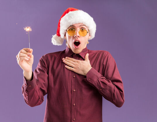 手令人印象深刻的年轻金发男子戴着圣诞帽和眼镜拿着节日火花看着相机保持手放在胸部隔离紫色背景胸前帽子看着