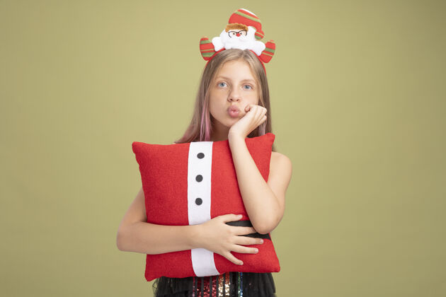 小小女孩穿着闪亮的派对礼服 戴着头巾 圣诞老人拿着一个有趣的枕头 手放在下巴上 站在绿色的墙上等待着圣诞节闪光下巴