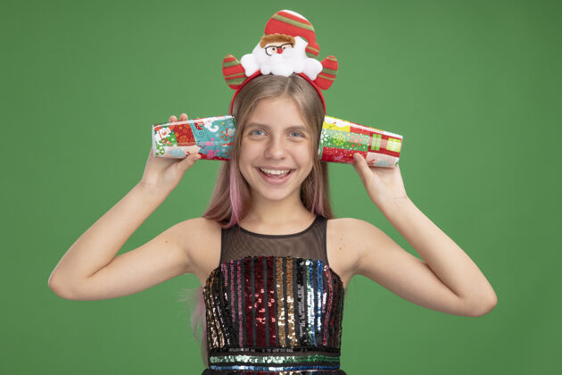 站着小女孩穿着闪亮的派对礼服 戴着圣诞老人的头巾 耳朵上戴着五颜六色的纸杯 站在绿色的墙上 开心地笑着圣诞节圣诞老人小