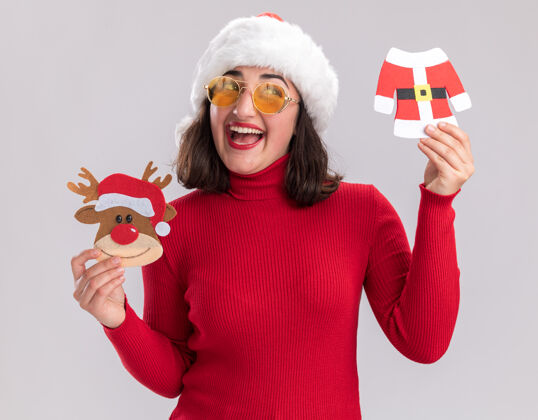 站着快乐的小女孩穿着红毛衣 戴着圣诞帽 戴着眼镜 拿着圣诞玩具 面带微笑地站在白色的墙上 望着一旁微笑年轻人圣诞节