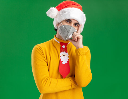 表情穿着黄色高领毛衣 戴着圣诞老人帽 打着滑稽领带 戴着面罩 手放在下巴上 严肃地看着镜头 站在绿色背景下思考高领毛衣下巴严肃