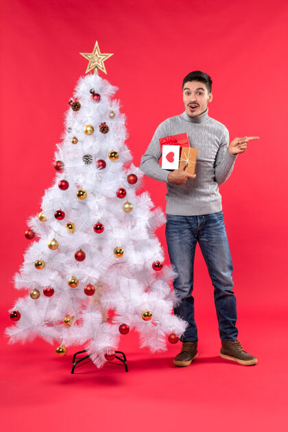 圣诞节困惑的帅哥站在装饰过的白色圣诞树旁 手里拿着他的礼物离开迷茫站着
