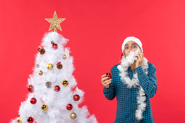圣诞一个戴着圣诞老人帽子 穿着蓝色条纹衬衫 手里拿着装饰品的年轻人站着圣诞老人配件圣诞树