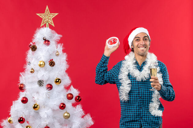 站着滑稽的年轻人戴着圣诞老人的帽子 举起一杯葡萄酒 手里拿着一个挂钟 站在红色的圣诞树旁年轻人圣诞老人酒