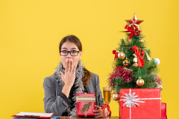 嘴前视图惊讶的女孩戴着眼镜坐在桌旁 把手放在嘴边圣诞树和礼物鸡尾酒惊讶女孩鸡尾酒礼物