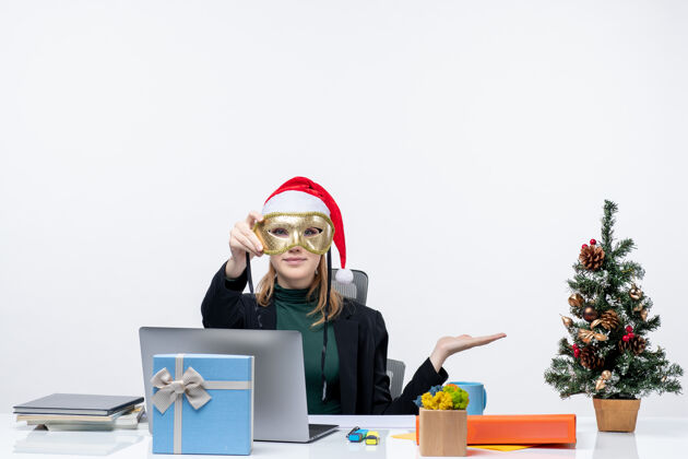 面具戴着圣诞老人帽子和面具的年轻女子坐在桌子旁 手里拿着圣诞树和礼物办公室工作场所年轻女人