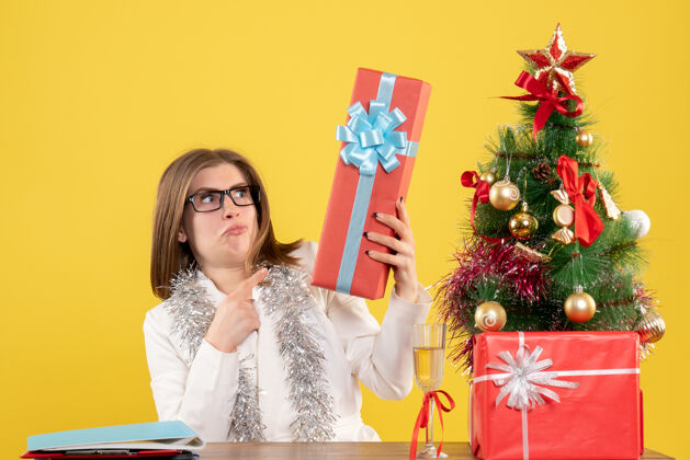 坐着正面图：女医生坐在桌子前 拿着礼物 黄色背景上的圣诞树和礼盒前面背景女医生
