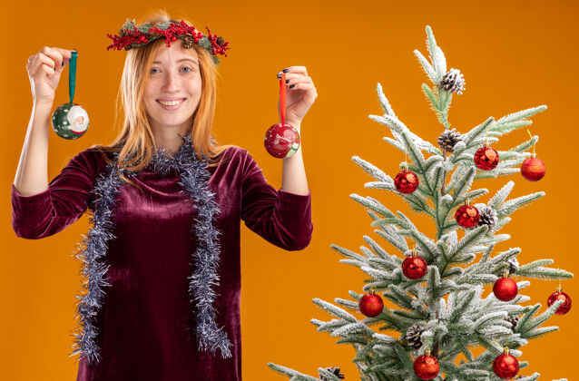 舞会微笑着站在圣诞树旁的年轻漂亮女孩身穿红色连衣裙 脖子上戴着花环 手里拿着橙色背景上孤立的圣诞球衣服抱着脖子