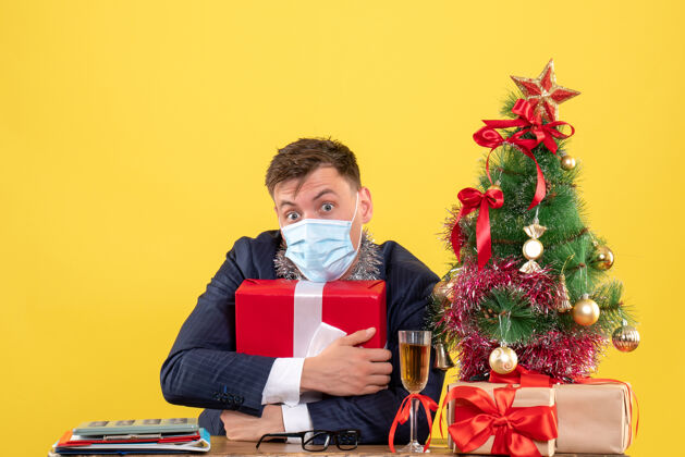 蝴蝶结正面图：商人紧紧地拿着礼物坐在圣诞树旁的桌子旁 黄色的礼物桌子领带商务