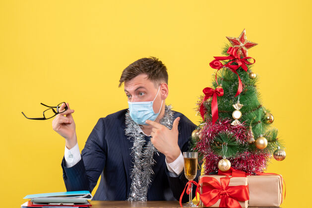 树前视图中的商人指着他的面具坐在圣诞树旁的桌子和黄色的礼物男人面具生意人