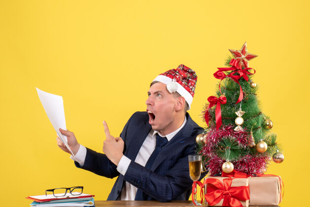 前面愤怒的男人展示文件坐在圣诞树附近的桌子上 黄色墙壁上的免费空间礼物的正面视图西装礼物快乐
