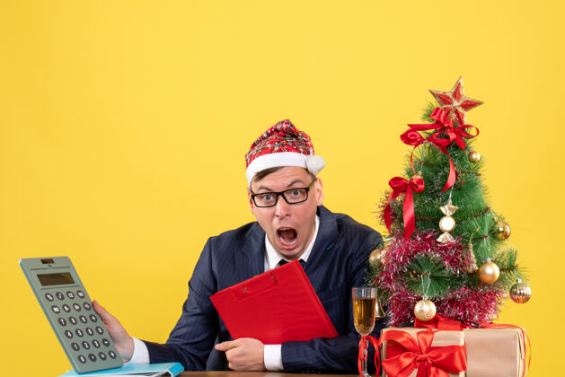 套装商务人士手持计算器坐在圣诞树旁的桌子前 呈现黄色桌子抱着成人