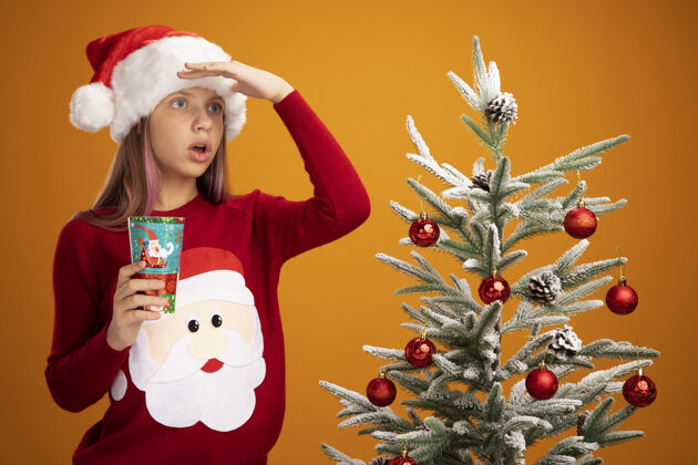 树小女孩穿着圣诞毛衣 戴着圣诞帽 手里拿着五颜六色的纸杯 远远望着橙色背景下的圣诞树头纸圣诞老人