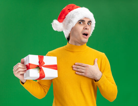 惊讶快乐的年轻人穿着黄色高领毛衣 戴着圣诞老人帽 打着滑稽的领带 拿着礼物 站在绿色的背景上惊讶地看着相机年轻高领毛衣抱着