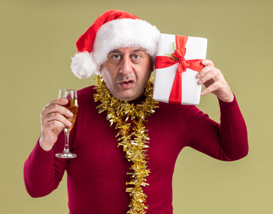 脖子中年男子戴着圣诞老人帽子 脖子上戴着金属丝 手里拿着圣诞礼物 手里拿着一杯香槟 站在绿色背景上困惑地看着摄像机站着圣诞老人困惑