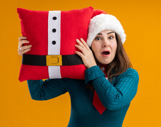 橙色焦虑的成年白人妇女 戴着圣诞帽 打着圣诞领带 拿着装饰好的枕头 孤立地放在橙色背景上 留着复制空间枕头圣诞老人成人