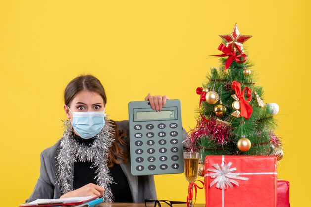 计算器正面图：戴着医用面罩的严肃女孩坐在桌子旁 手里拿着计算器圣诞树和鸡尾酒礼物坐着电话圣诞节