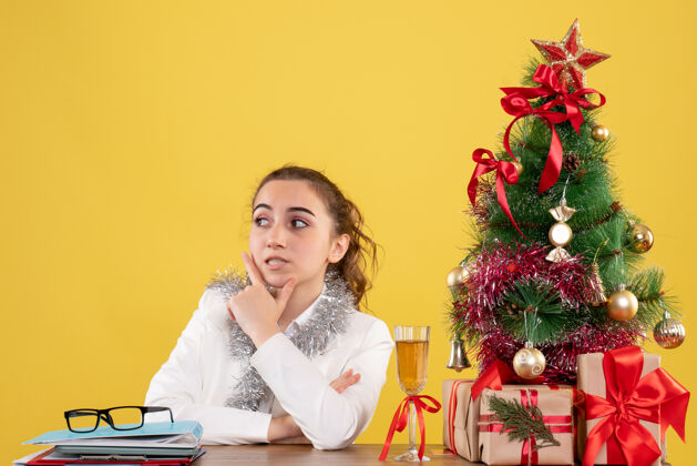 圣诞节正面图：女医生坐在黄色背景的桌子后面 还有圣诞树和礼品盒插花微笑插花