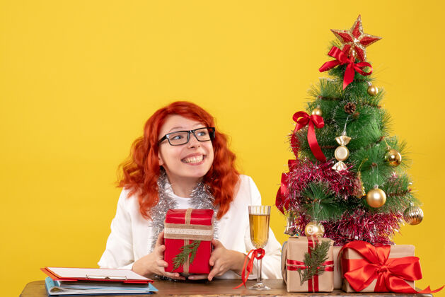 圣诞节前视图：女医生坐在桌子后面 黄色背景上有圣诞礼物安排礼物前面