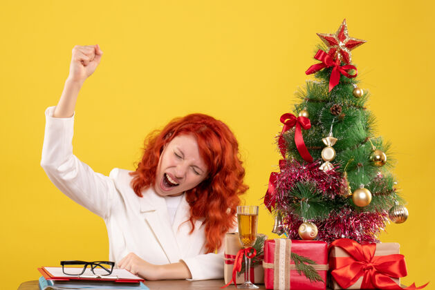 人正面图：女医生坐在桌子后面 拿着圣诞礼物 黄色背景上的树前面健康背景