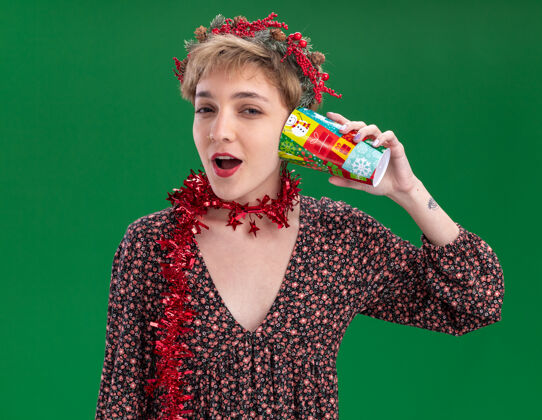 漂亮好奇的年轻漂亮女孩戴着圣诞花环 脖子上戴着金属丝花环 耳边拿着塑料圣诞杯 听着隔离在绿墙上的秘密听花环花环