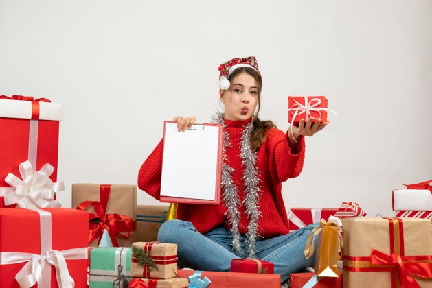 快乐戴着圣诞帽的快乐女孩拿着礼物和文件坐在白色的礼物旁边盒子女孩到处